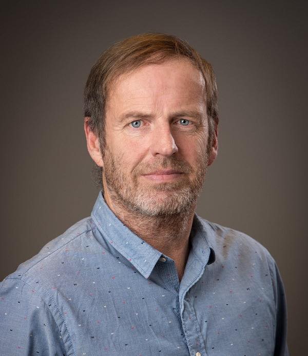 Mann i blå skjorte foran mørkgrå bakgrunn